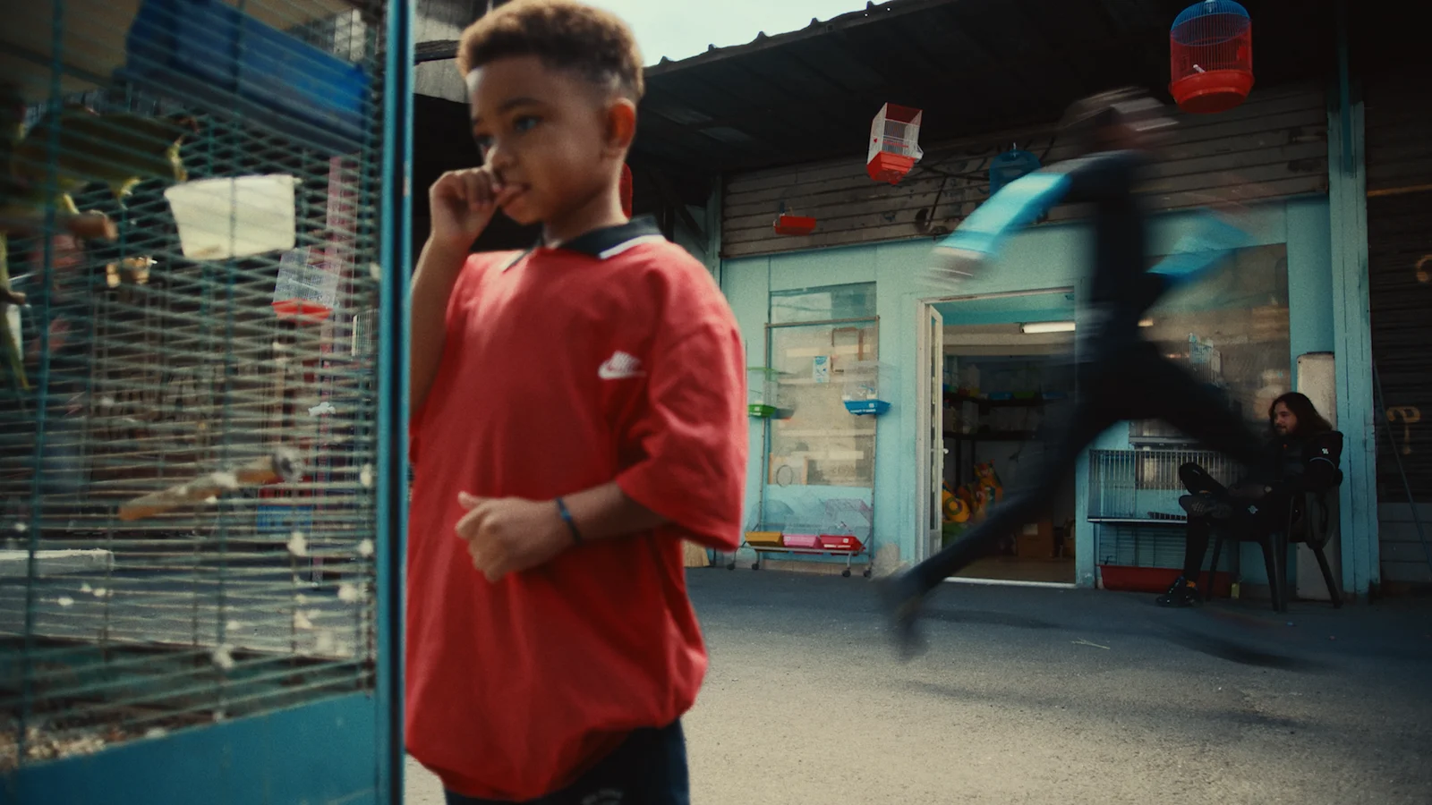 Young child in a red shirt looks at a bird cage outside a small shop while a person runs past in motion blur