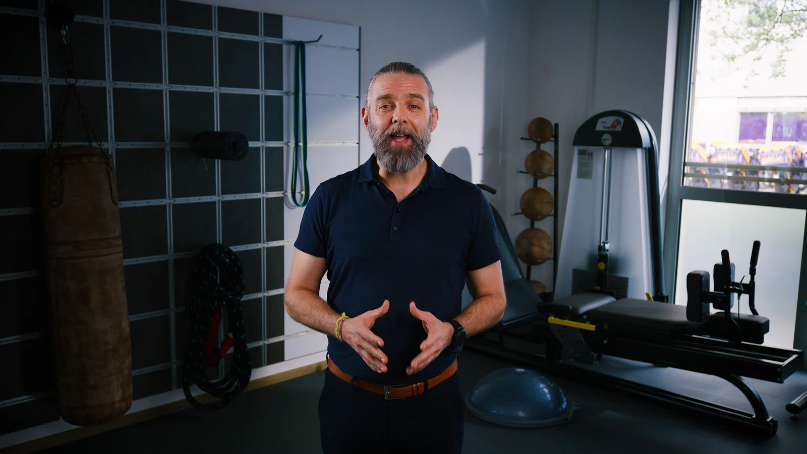Middle aged fitness coach speaks in a gym with a punching bag and exercise equipment behind him