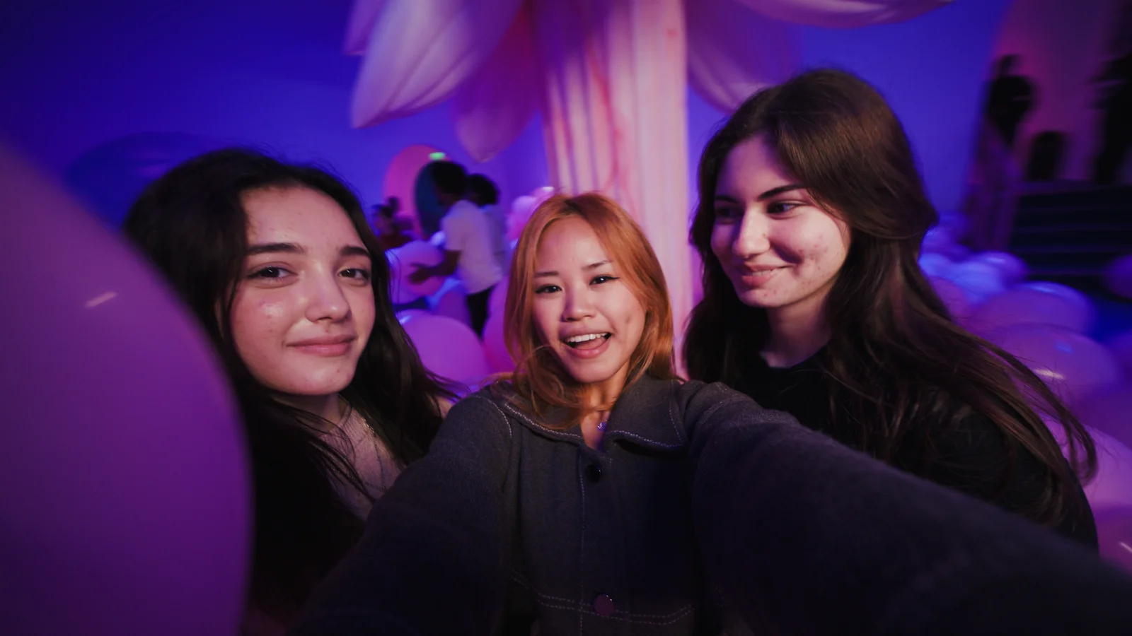 Three girls pose for a selfie, smiling in a moody purple-pink room with a huge pink palm and children playing with pink balloons in the background