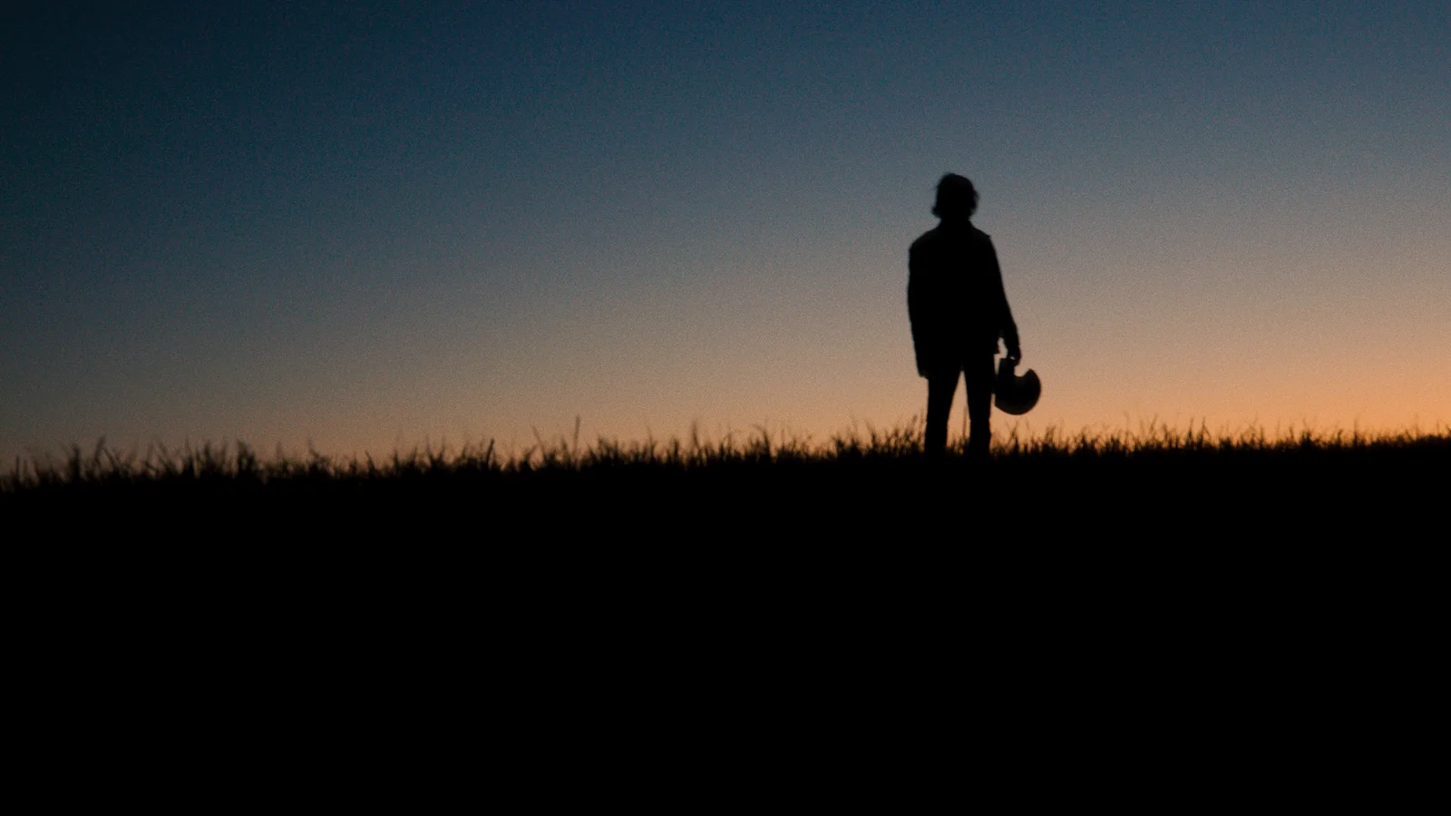 A silhouetted figure of a man stands on the horizon, watching the sunset fade away and holding a motorcycle helmet